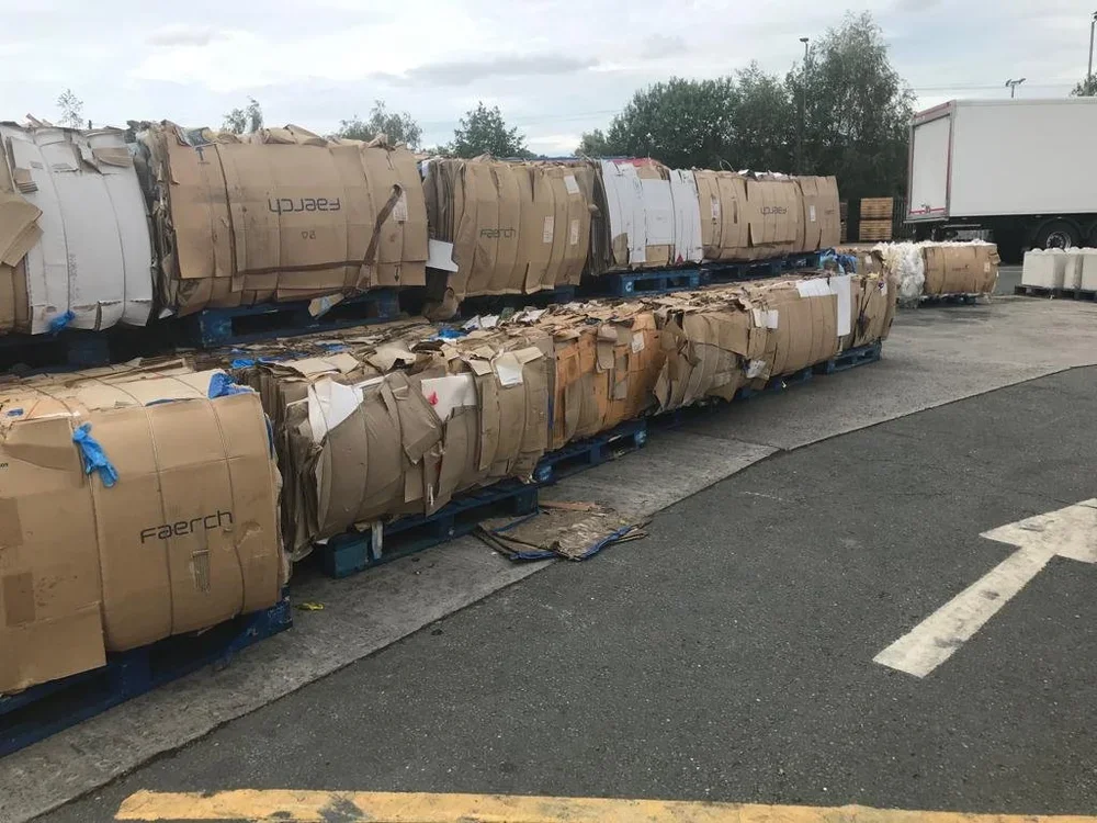 Piles of large flat cardboard boxes and paper packaging stacked on blue pallets outdoors near a parking lot, with a white truck visible in the background.