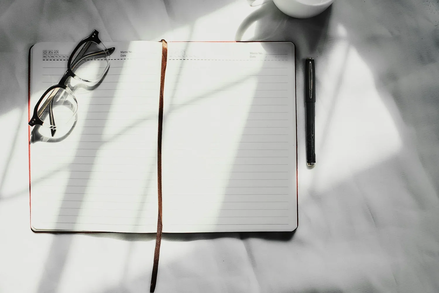 Open notebook with lined pages, a pair of reading glasses on top, a black pen to the right, and a white coffee cup above, all on a white surface with shadows.