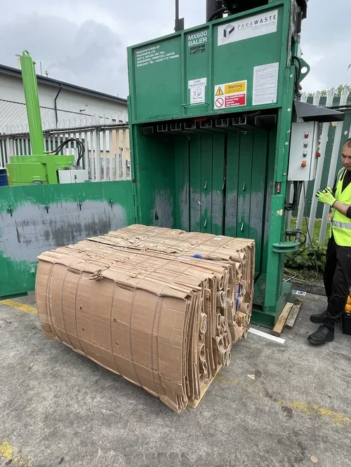 Large cardboard rolls loaded into a green industrial baler machine at a waste recycling facility. A worker in a yellow safety vest and gloves stands nearby.