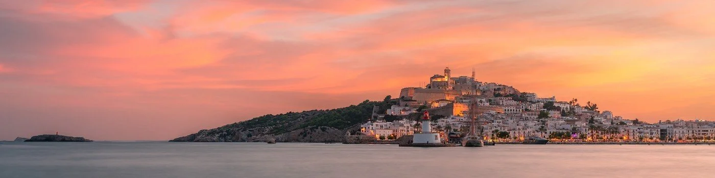 Sunset over a coastal town with white buildings on a hill, a lighthouse near the shore, and calm waters in the foreground.