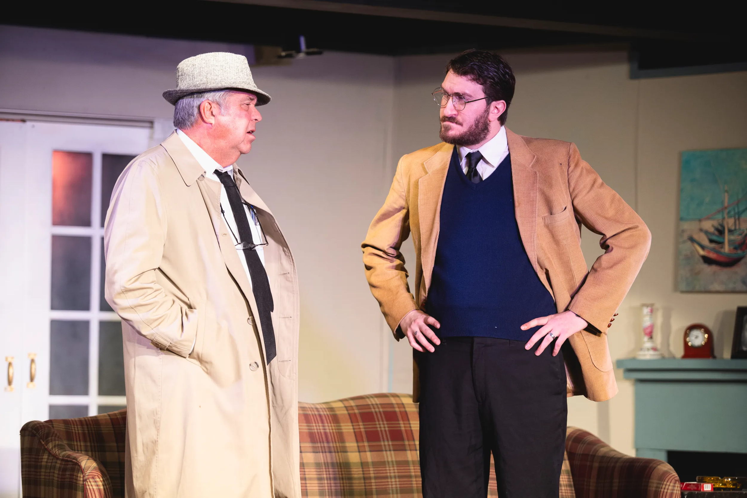 Two men having a serious conversation in a living room, one wearing a beige trench coat and hat, the other in a brown blazer and glasses.