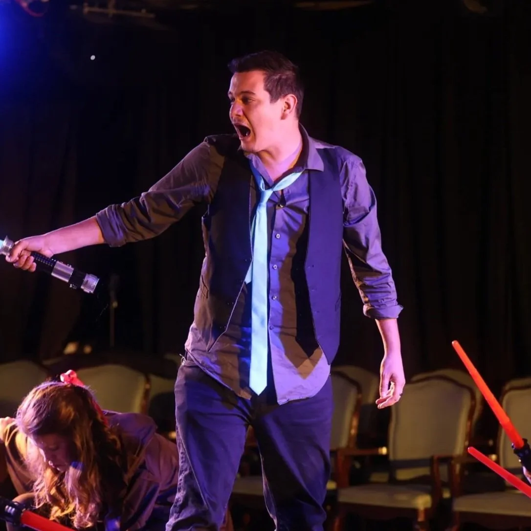 A man on stage, dressed in a purple shirt with rolled-up sleeves, black vest, and loosened tie, appears to be shouting or singing passionately, holding a flashlight in his right hand. A woman with long reddish hair and a hair clip is kneeling on the stage in front of him, seemingly engaged in a theatrical performance. The background is dark with empty chairs, suggesting a theater setting.