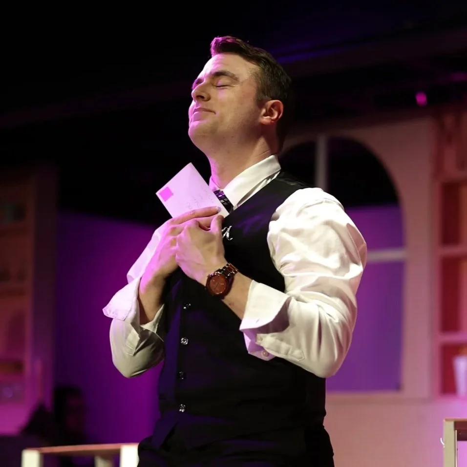 A man in formal attire, with eyes closed and a content smile, holding a small white card close to his chest, standing in a room with purple and black lighting.