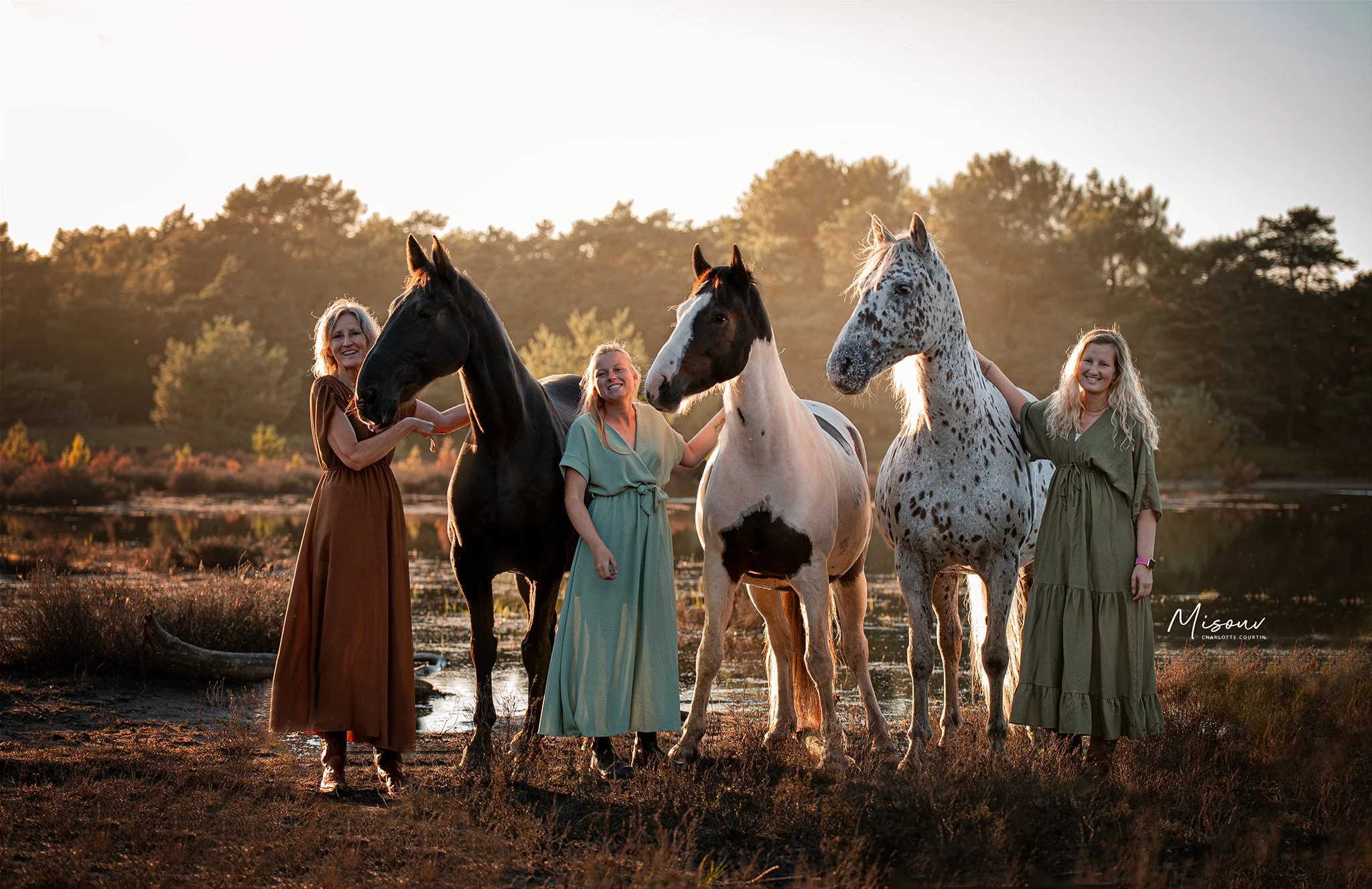 Three women standing in a field with three horses; one woman wears a brown dress, another a green dress, and the last a light blue dress. The horses are black, black and white, and white with spots.