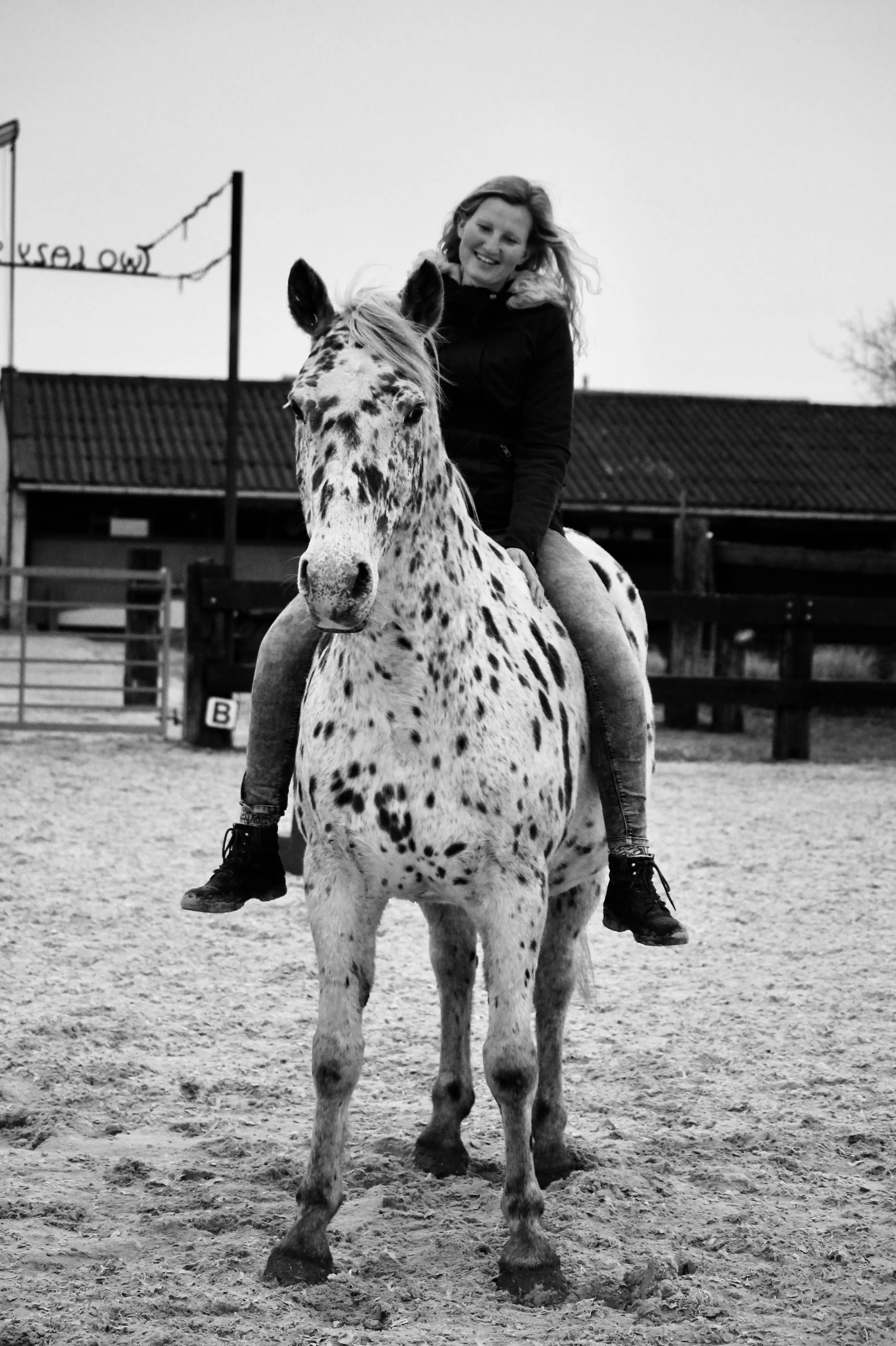 Black and white photo of a person riding a spotted horse in an outdoor setting.