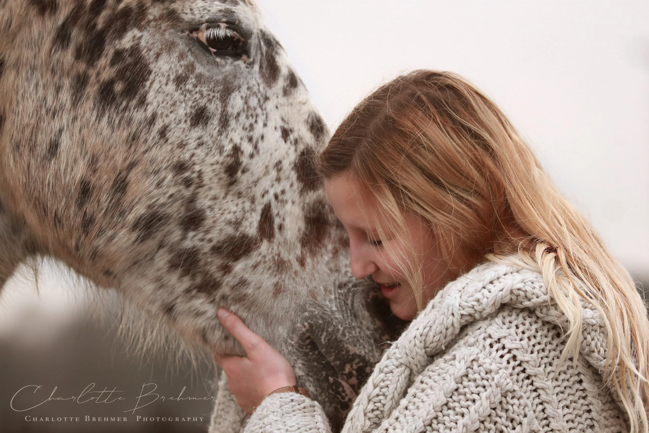 Woman gently nuzzling a spotted horse while wearing a knitted sweater.