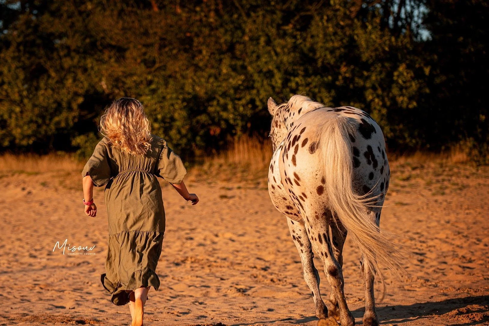Woman in green dress walking with a spotted horse on a sandy path, surrounded by trees.