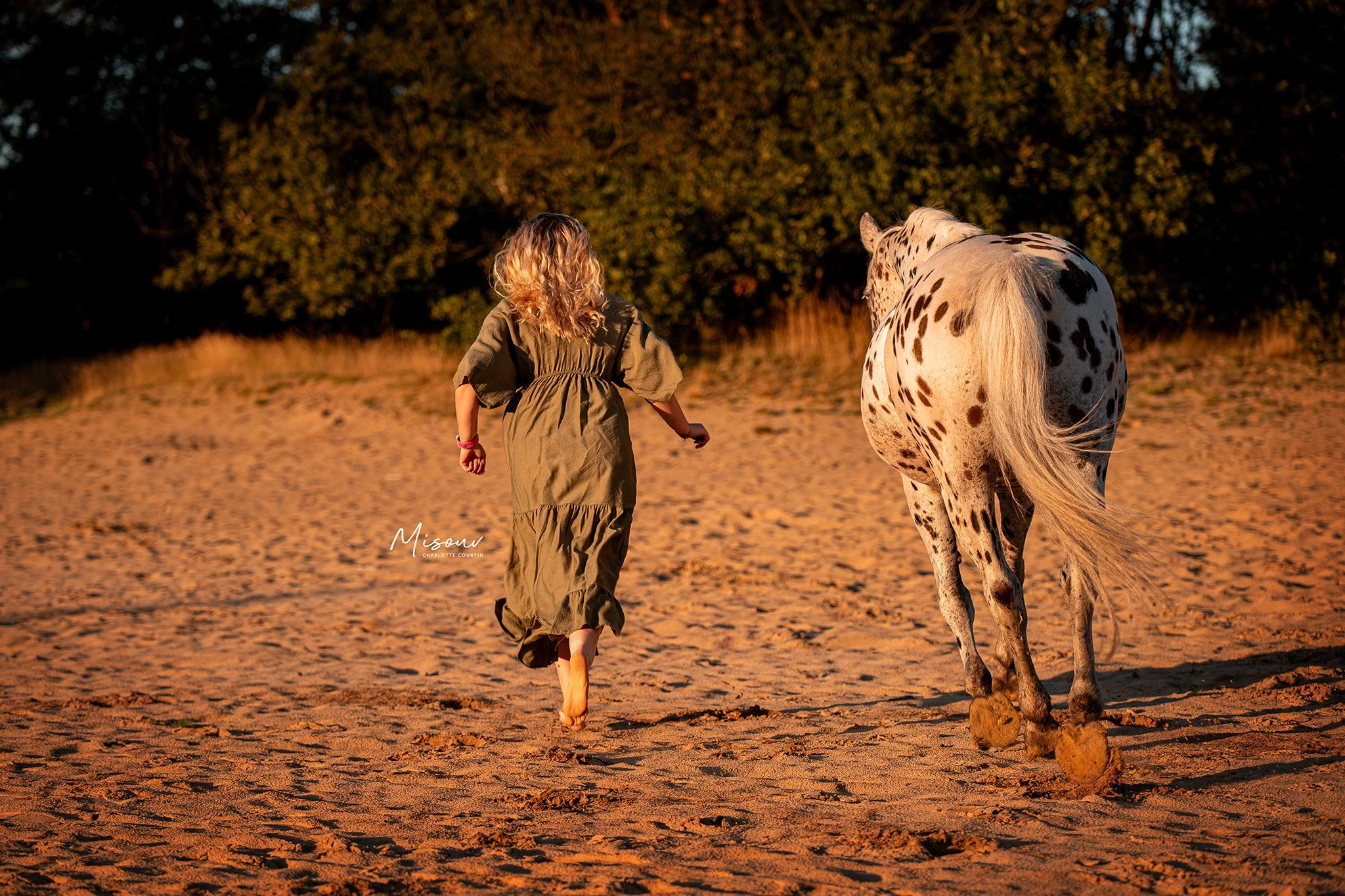A child in a green dress and a spotted horse walking on a sandy path at sunset with trees in the background.