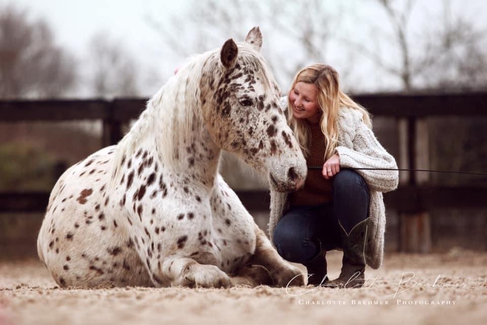 A woman crouching next to a speckled horse lying on the ground in a sandy area. The woman is wearing a cozy sweater and smiling at the horse.