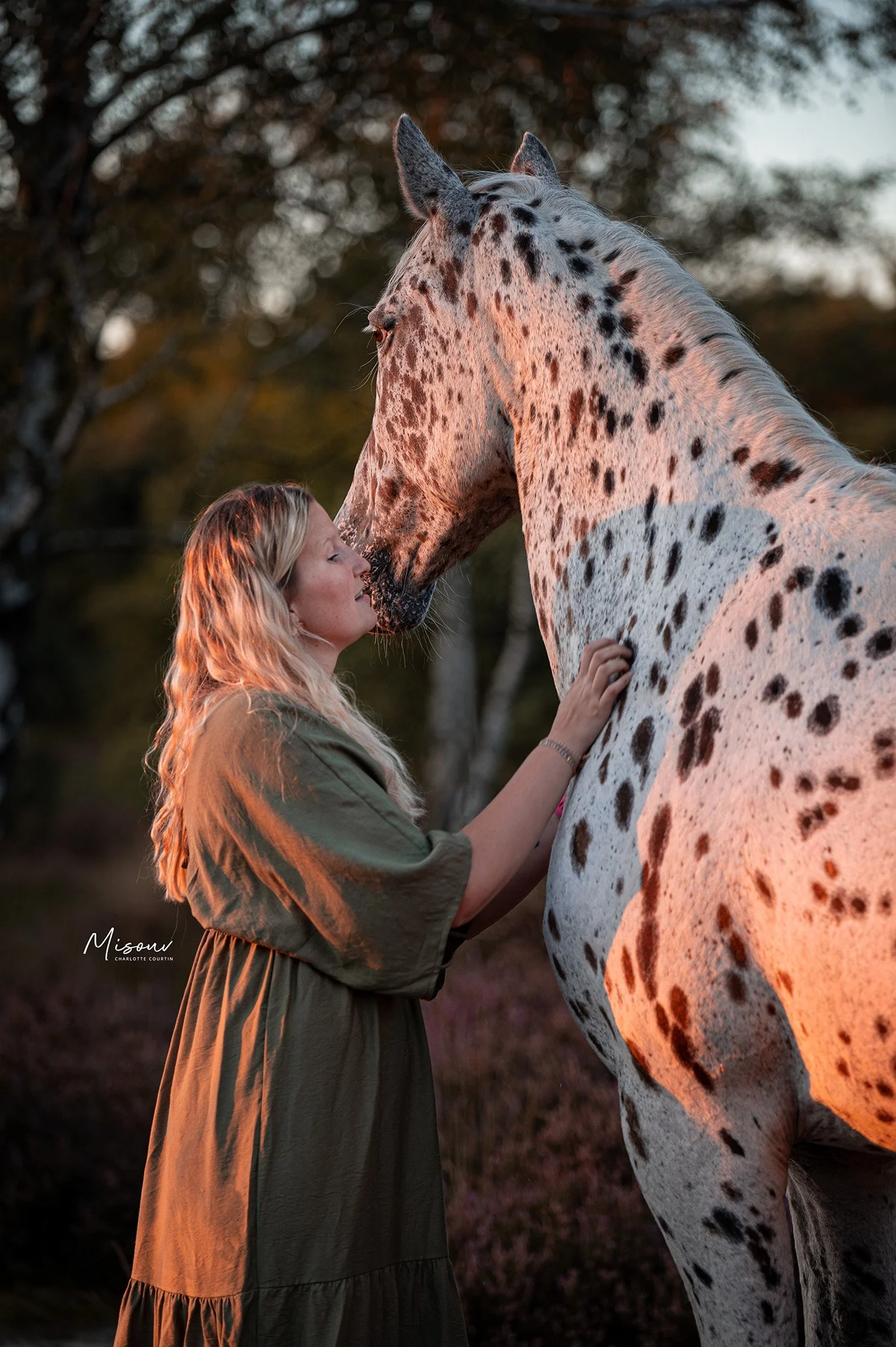 Woman in green dress gently caressing a spotted horse in a sunset-lit forest.
