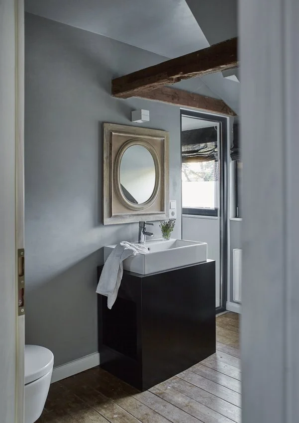 Ein modernes Badezimmer mit grauen Wänden getsrichen in dem Farbton fog aud der Farbkarte Morning MIst von der Marke Paint of Walinoon, einem weißen Waschtisch mit schwarzem Unterschrank, einem ovalen Spiegel, einem Fenster und einer Holzdecke.
