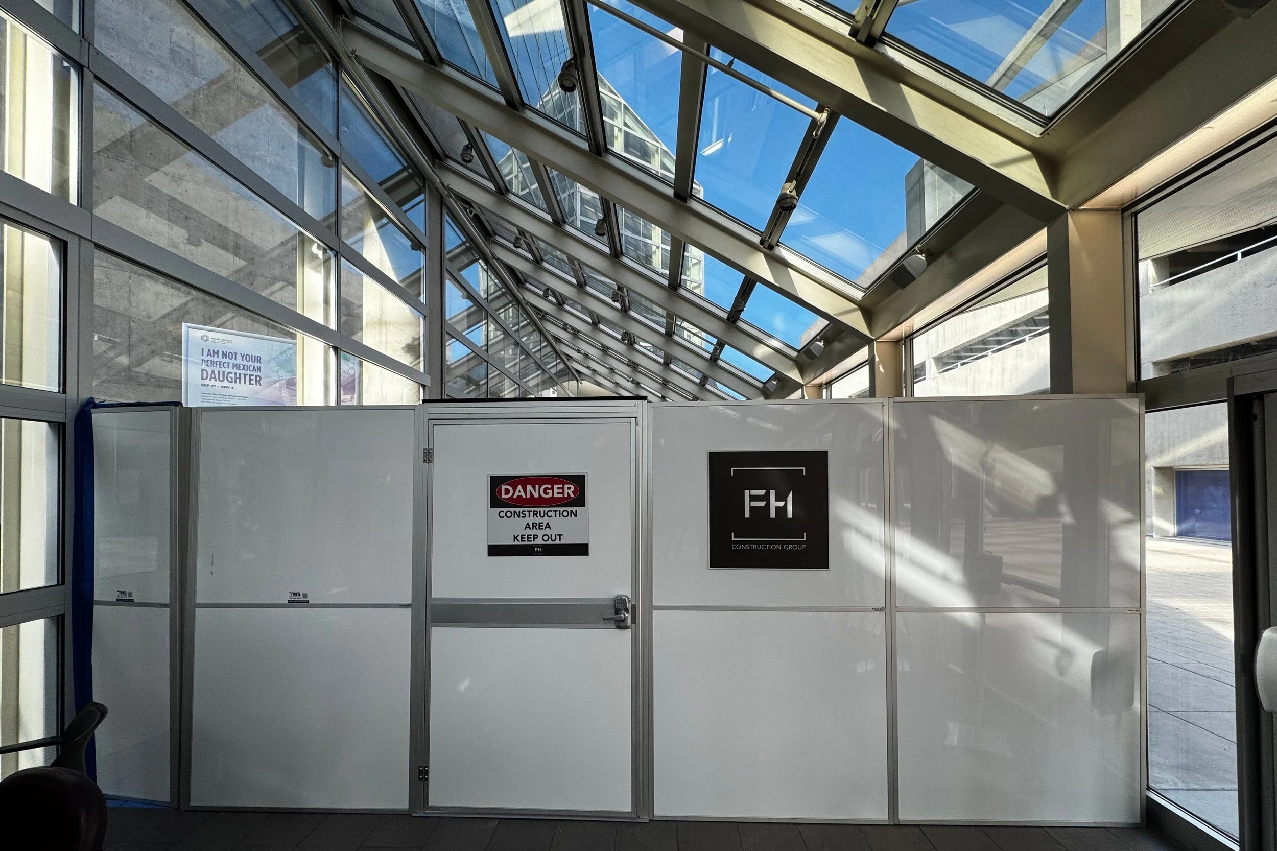 Construction barricade inside a modern building with glass windows and ceiling, featuring warning signs that say 'Danger Construction Area Keep Out' and a logo for FHI Construction Group.