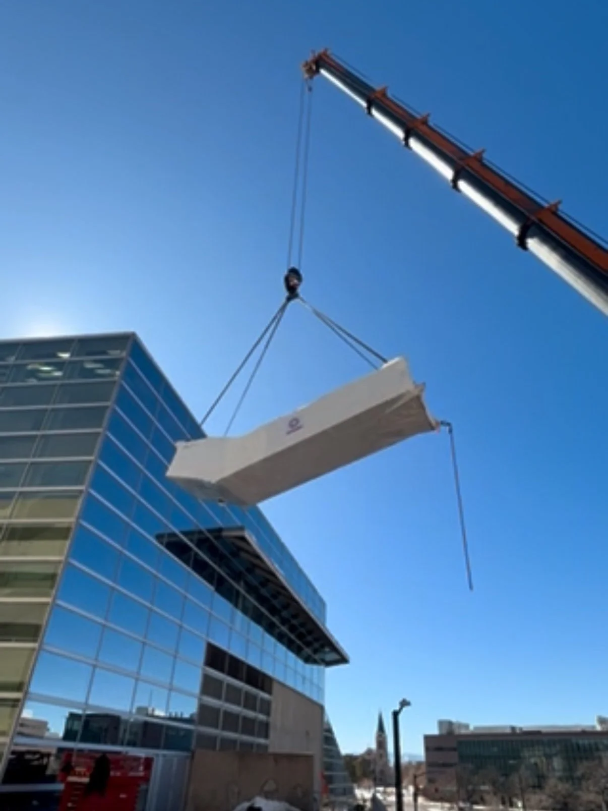 Construction crane lifting a large, white insulation panel onto a modern glass building under a clear blue sky.
