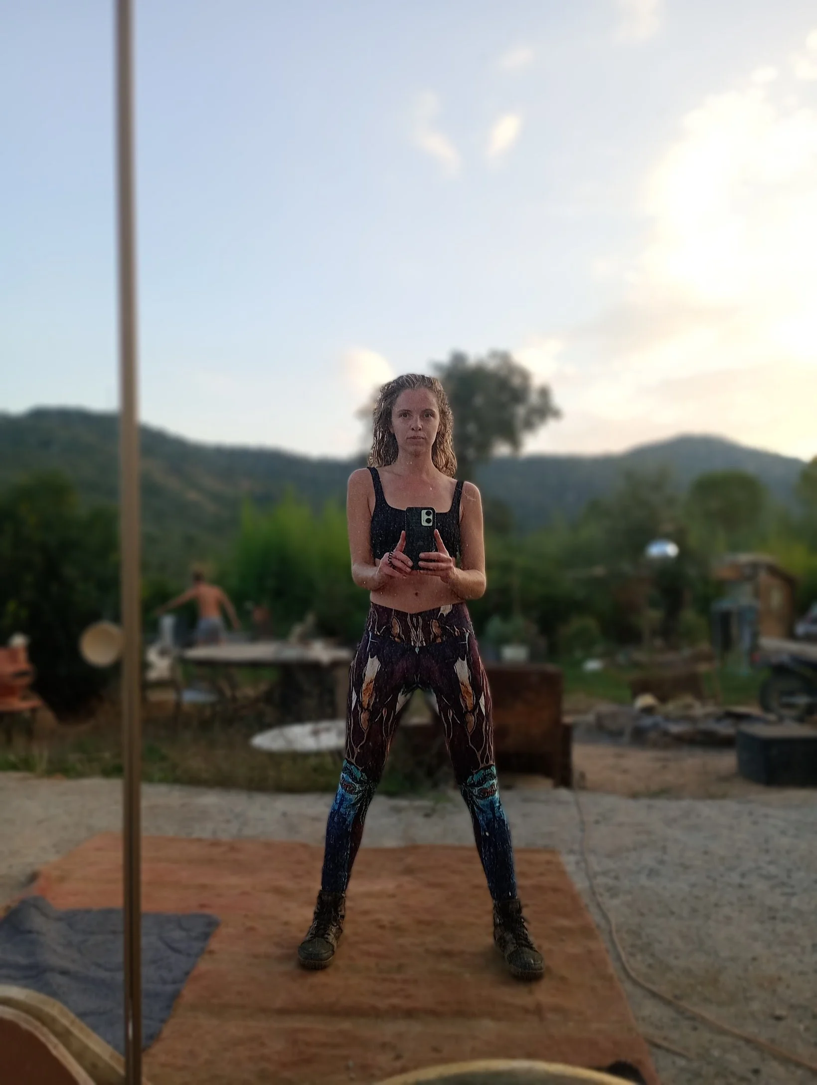 Jeune femme posant devant un miroir naturel, portant un haut noir et un pantalon à motifs colorés, avec un paysage de montagnes et de verdure en arrière-plan, au coucher du soleil.