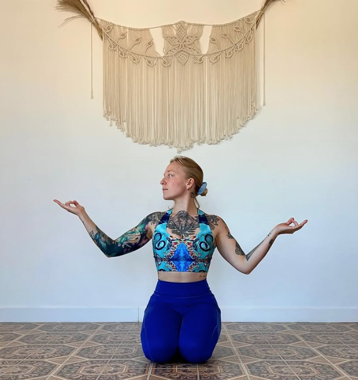 Femme tatouée en position de méditation ou yoga, à genoux sur un sol en carreaux, avec un motif boho: haut coloré et pantalon bleu, décorations en macramé au mur.