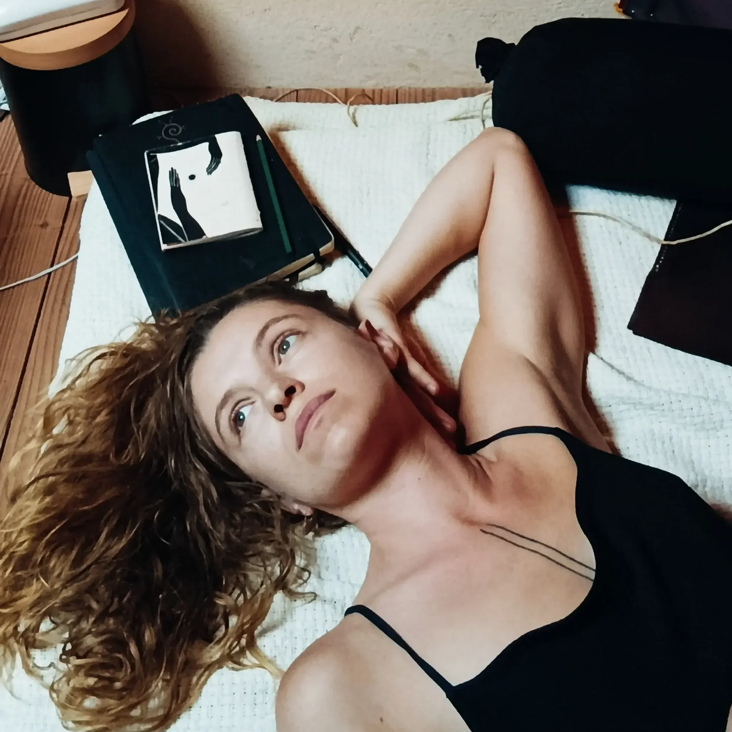 Une personne allongée sur un lit avec des cheveux roux bouclés, portant un haut noir, dans une pièce avec un mur en bois, avec un ordinateur portable sur le lit et un appareil médicinal à côté.
