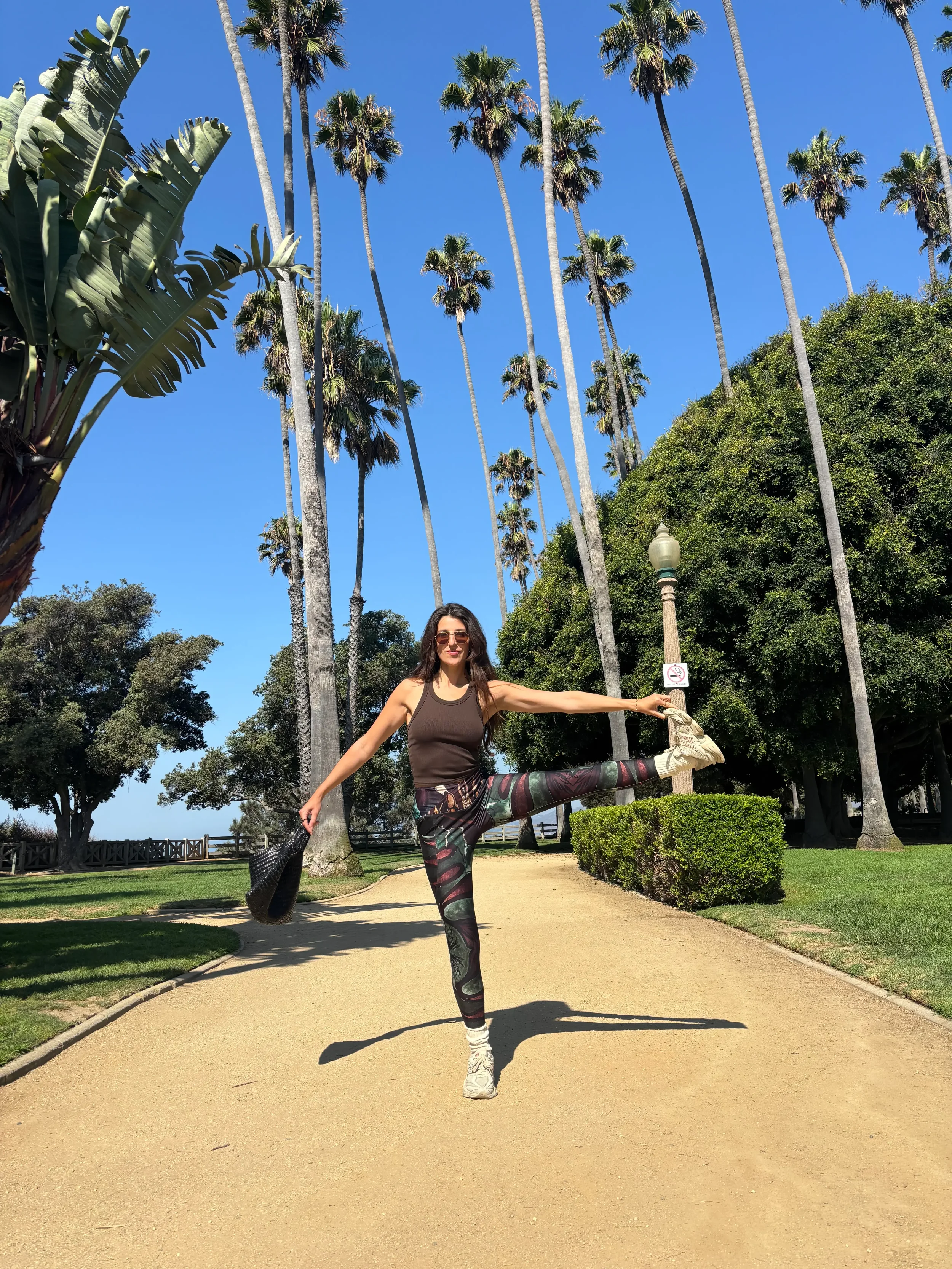 Art à porter - Verena Franzeti .Une femme fait une pose d'équilibre en étirant une jambe dans un parc avec des palmiers et un ciel bleu.