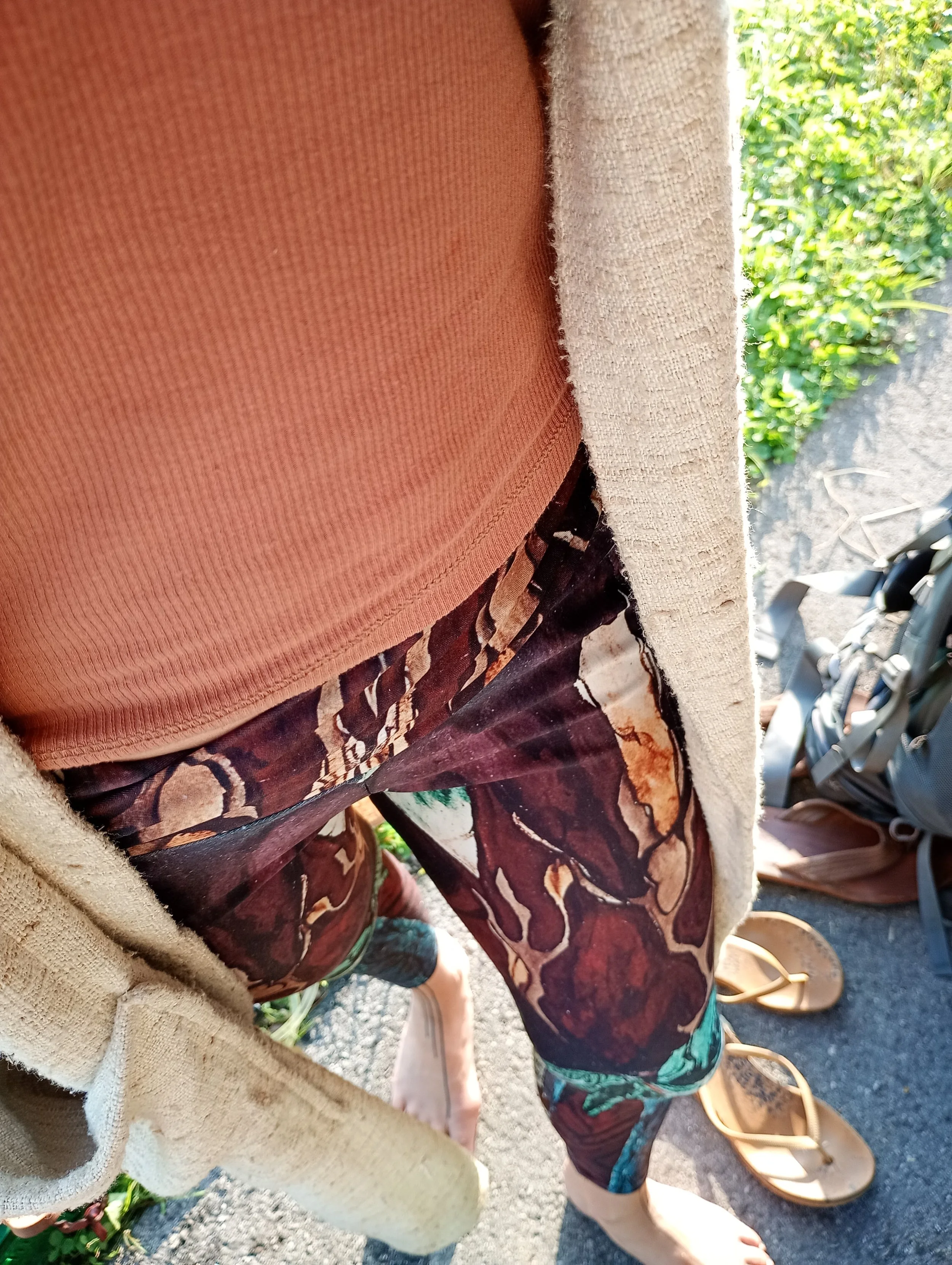 Une personne porte un t-shirt orange, une veste beige et un pantalon avec un motif marron, beige, vert et noir, en extérieur. Il y a des chaussures en cuir marron et des sandales sur le sol à côté d'une sacoche.