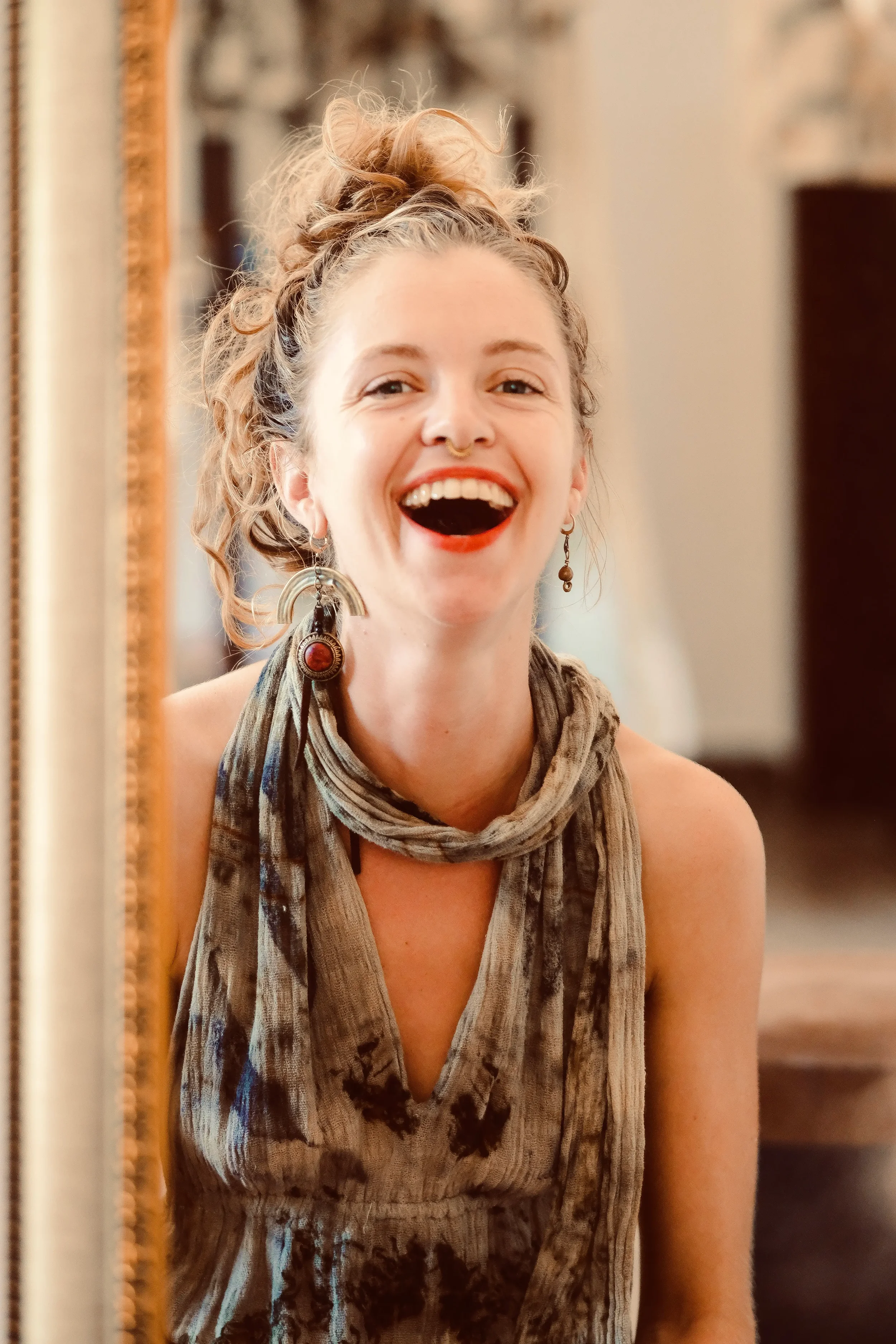 Verena Franzett jeune femme blonde éclatant de rire portant une robe, de longues boucles d'oreilles se regardant dans un miroir