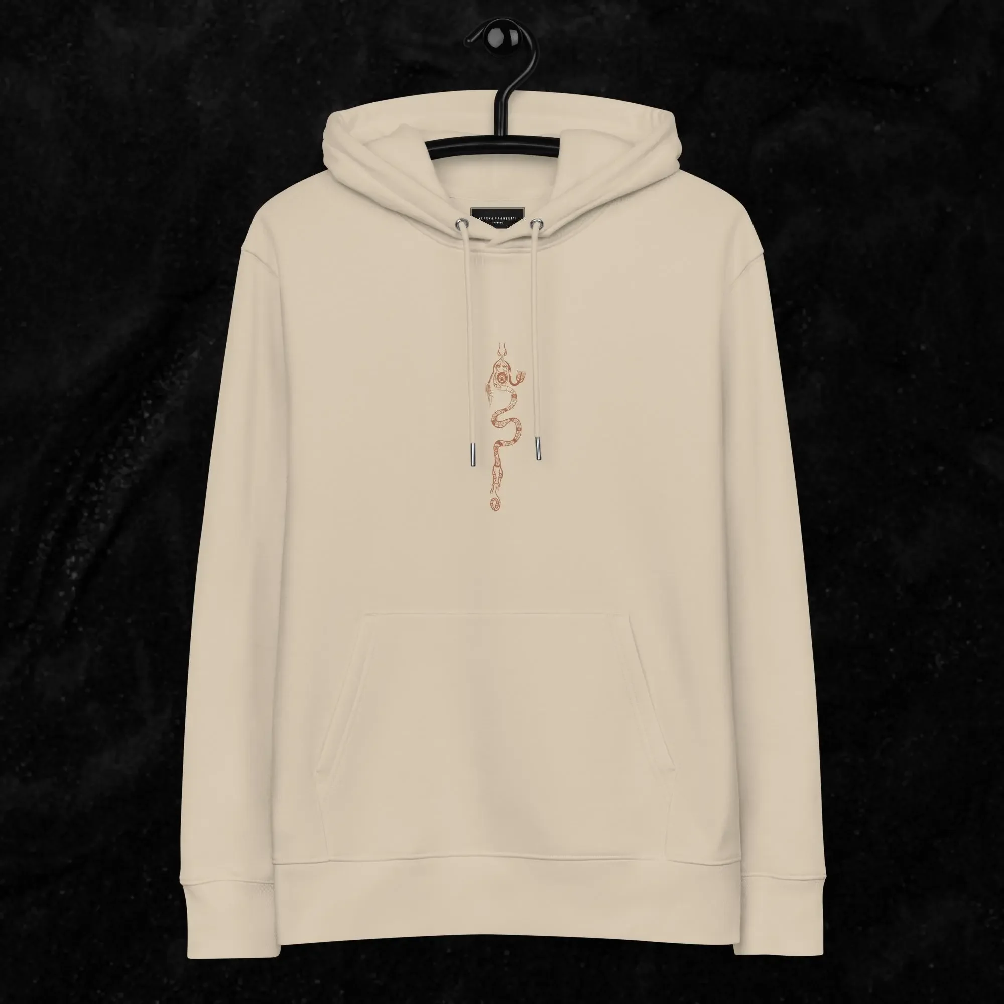Sweat à capuche beige avec un design tribal symétrique de couleur marron foncé, représentant une figure serpent organic, sur un fond noir. Verena Franzetti Collection Shape Shifting