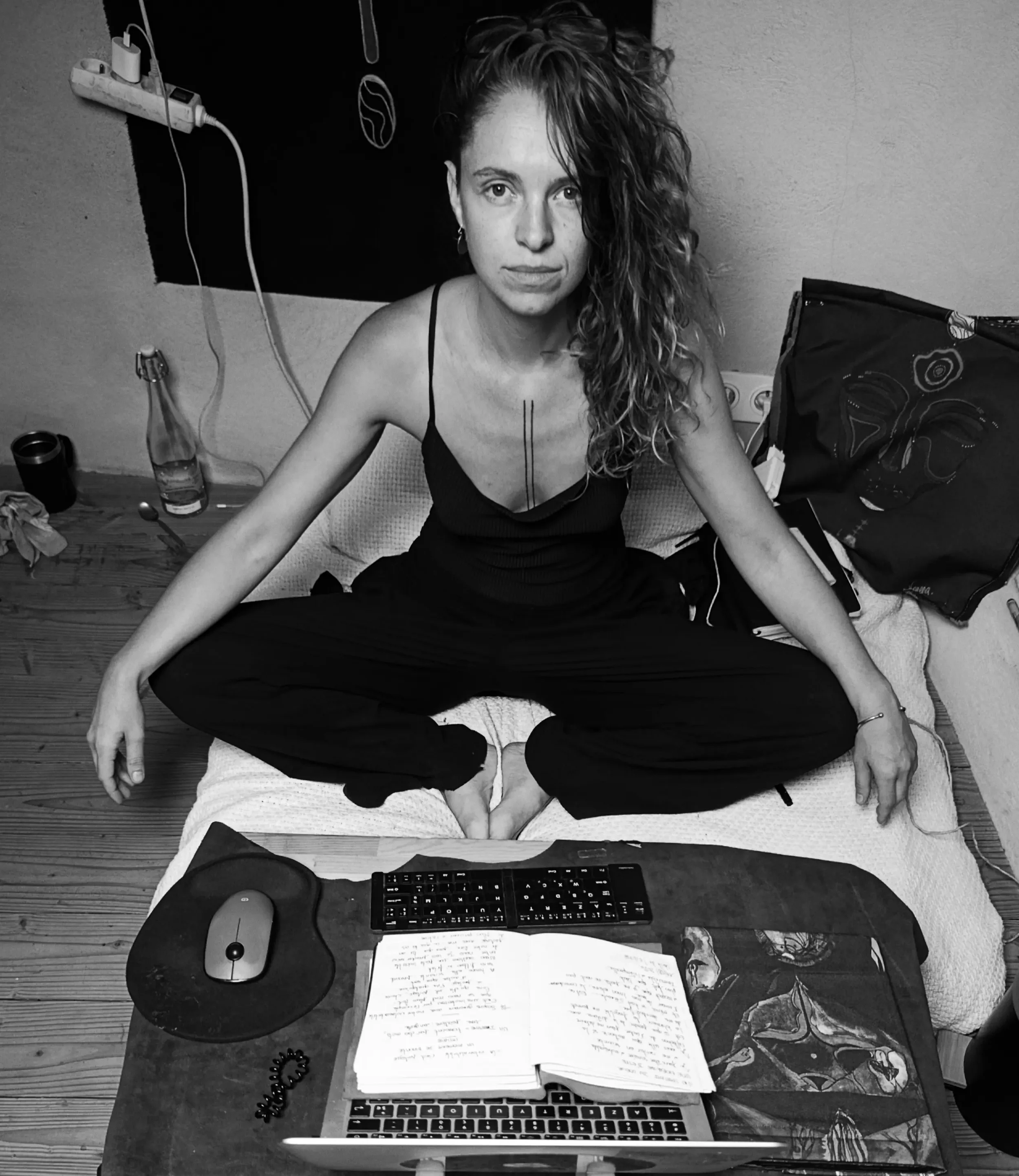 Un bureau de travail avec un ordinateur portable, une souris, un clavier, des notes manuscrites, un livre, des écouteurs et une bouteille en verre. En arrière-plan, une personne assise en position de méditation ou de yoga, les jambes croisées, dans une pièce avec un mur en papier peint.