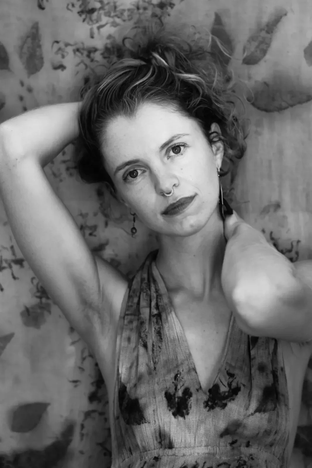 
Portrait en noir et blanc de Verena Franzetti avec des cheveux bouclés relevés, portant des boucles d'oreilles pendantes  adossée contre un mur avec des motifs vegetaux 