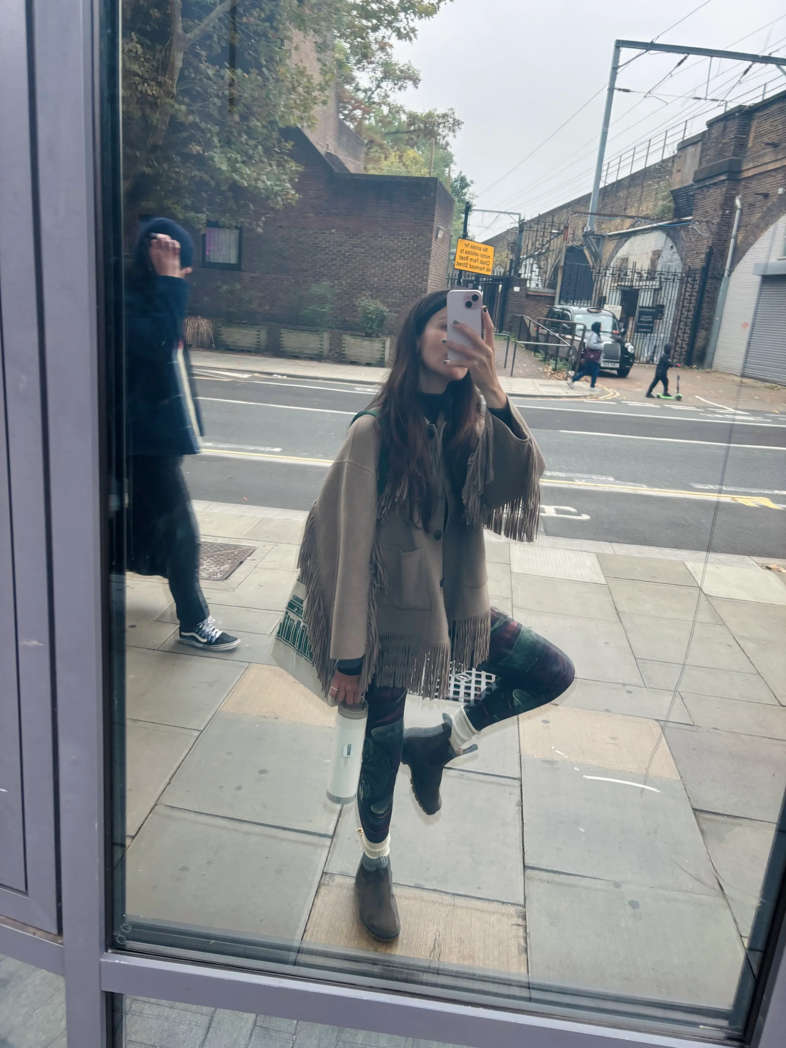 Art à porter - Verena Franzeti .Jessica prenant une photo d'elle-même dans une vitre d'une boutique, portant un manteau à franges, un legging au motifs organic de foret  des bottines noires, et tenant une bouteille blanche. On aperçoit également une 