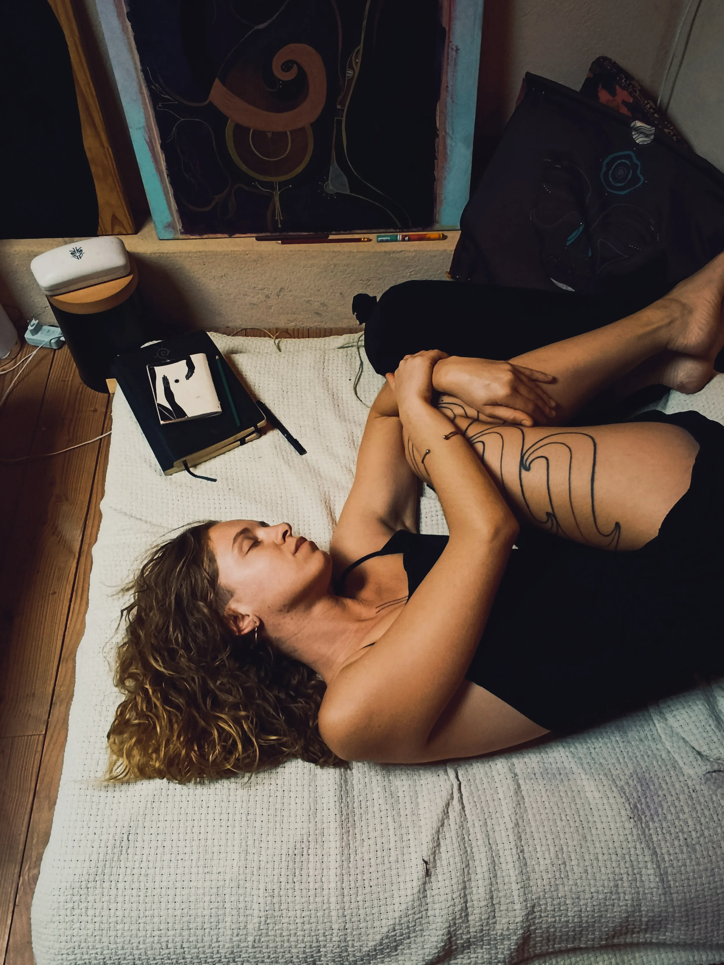 Une femme aux cheveux bouclés allongée sur un matelas blanc, les yeux fermés, avec un tatouage sur la jambe, tenant ses jambes avec ses mains, dans une pièce avec un tableau ou une peinture abstraite en arrière-plan.