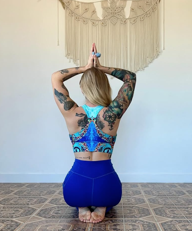 Une femme pratique le yoga en position de prière, vue de dos, avec des tatouages sur les bras, portant un haut coloré et un pantalon bleu, dans une pièce avec un tapis en bois et un macramé suspendu au mur.