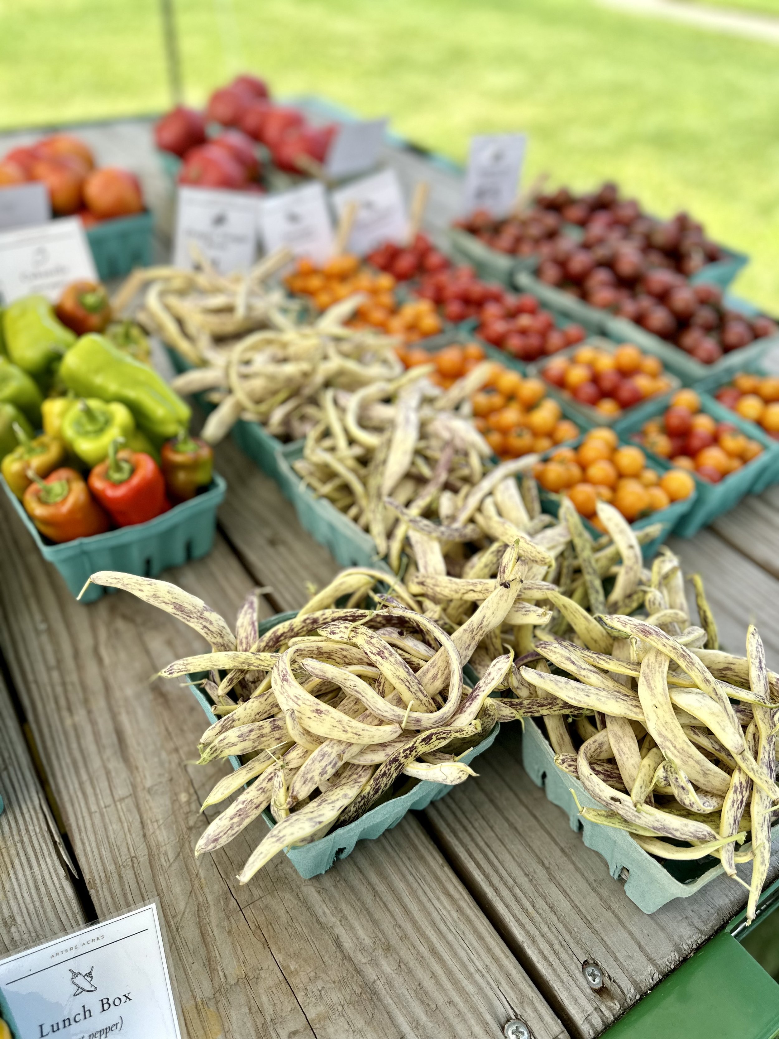 Assorted fresh vegetables on a wooden table, including yellow beans, mixed cherry tomatoes, bell peppers, and other produce in green cartons at a farmers market.