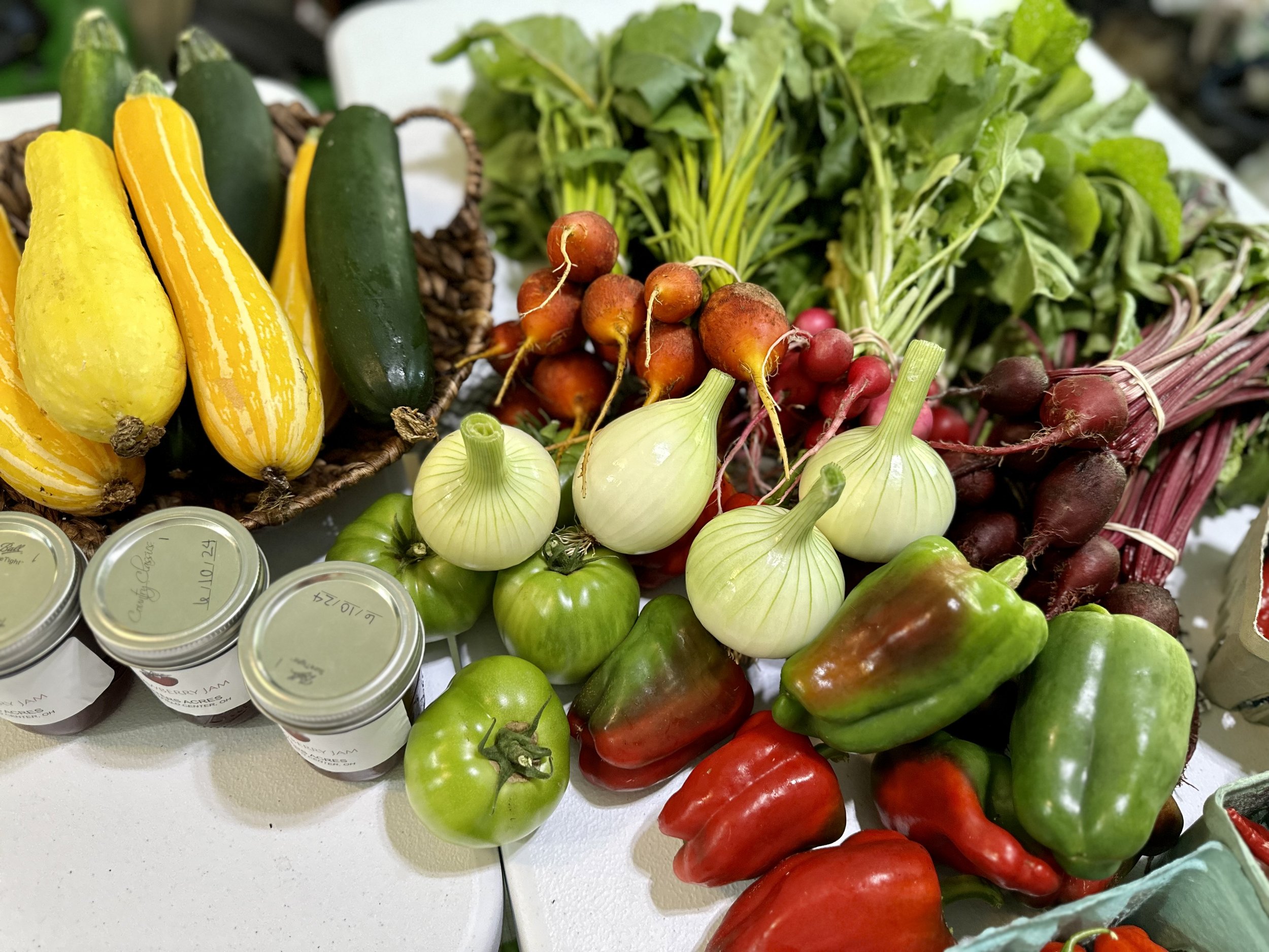 Assorted fresh vegetables including yellow squash, zucchini, beets, radishes, green tomatoes, onions, and bell peppers on a table with jars of preserves.