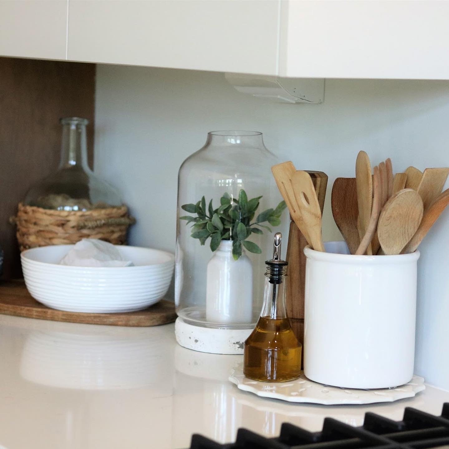 Kitchen countertop with a white ceramic utensil holder containing wooden cooking utensils, a glass bottle of olive oil, a glass cloche with decorative greenery, a white bowl, and a wicker basket with a large glass bottle inside.