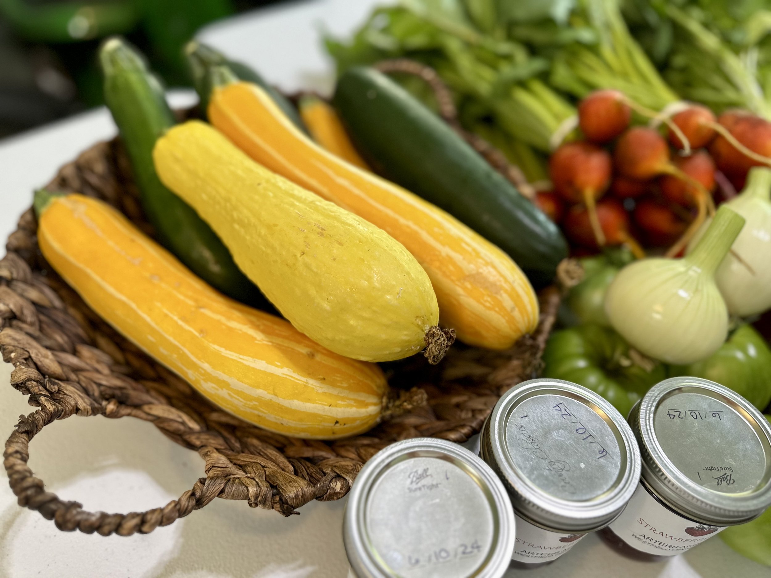 Basket with yellow and green squash, cucumbers, onions, radishes, and canned goods on a table.