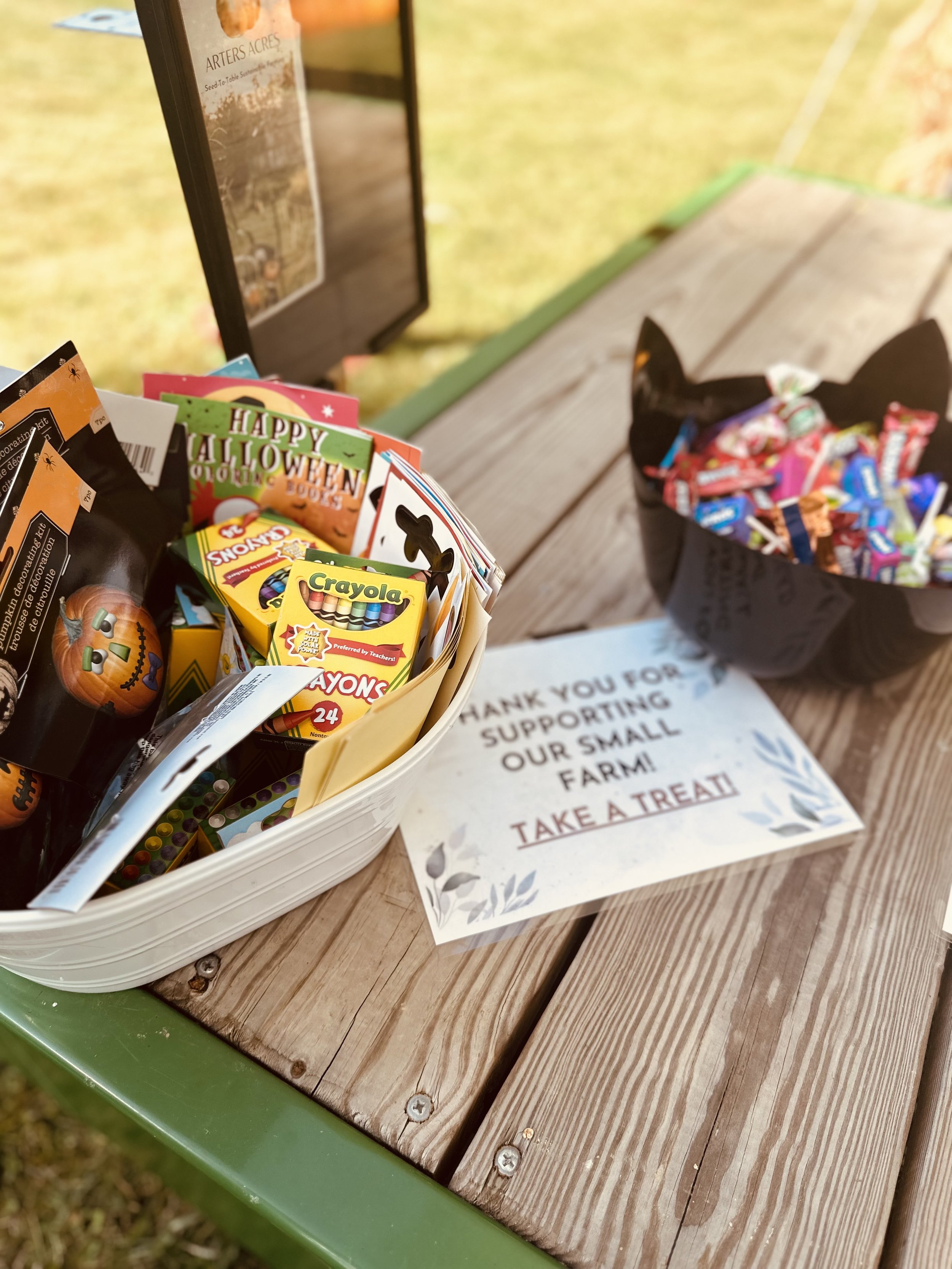 A table display with a basket containing Halloween-themed items including coloring books, crayons, and other small items. Next to it, there's a black container shaped like a cat filled with assorted candies. A sign reads "Thank you for supporting our