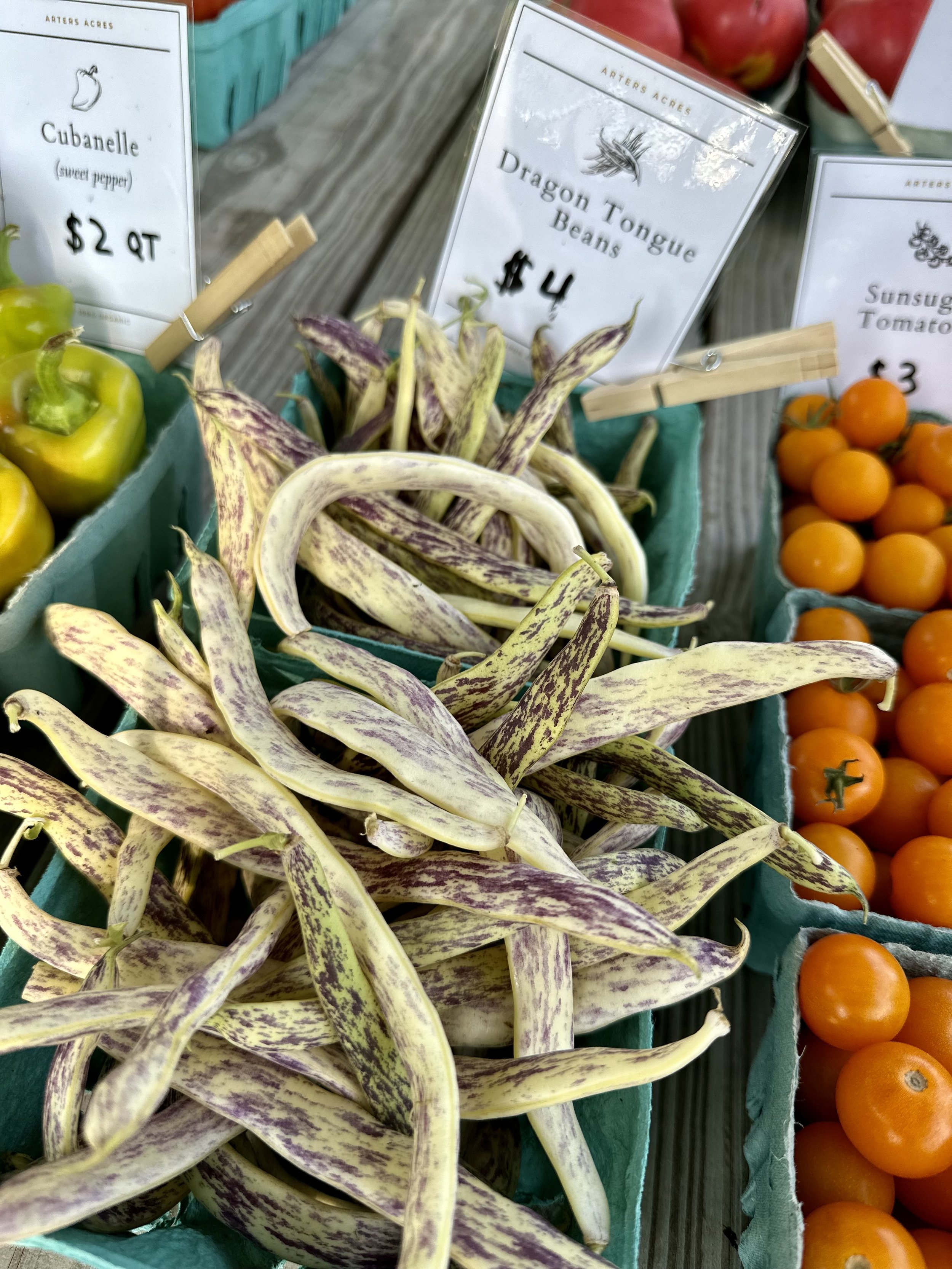Fresh produce display featuring Dragon Tongue Beans, Cubanelle peppers, and SunSugar tomatoes in teal containers with price labels at a market.