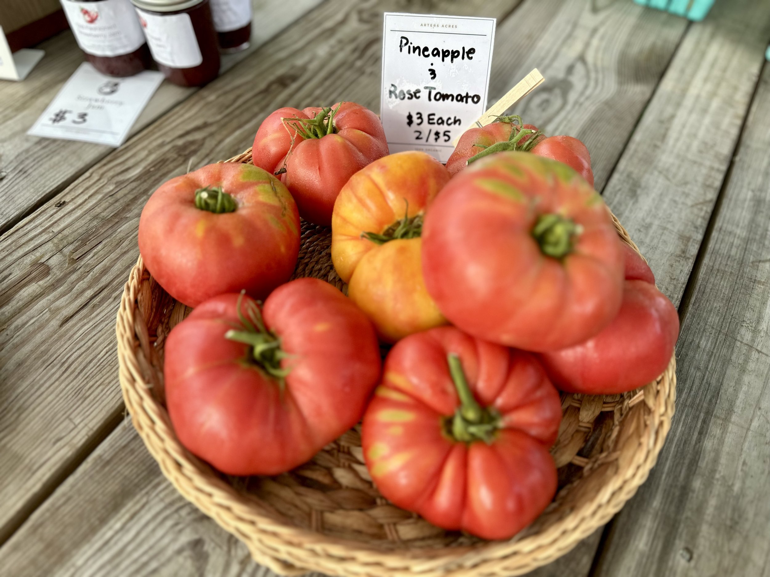 Basket of heirloom tomatoes labeled "Pineapple & Rose Tomato," priced at $3 each or 2 for $5, displayed on a wooden table.