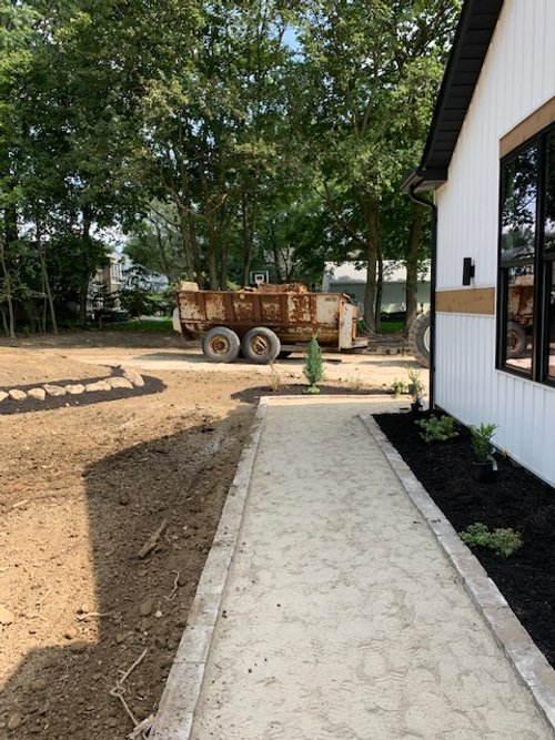 Newly constructed concrete pathway beside a modern building with a garden. An old, rusty trailer sits on dirt in the background, surrounded by trees.