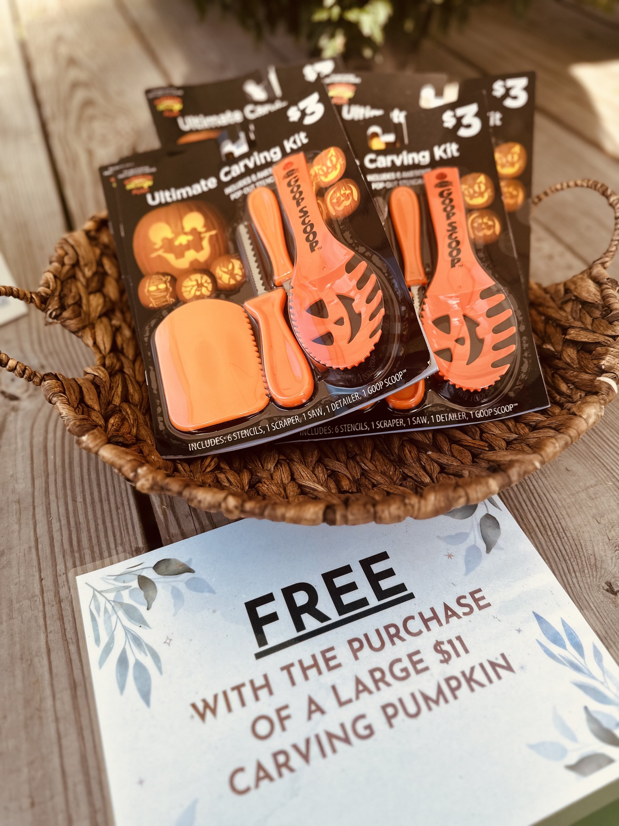 Pumpkin carving kits in a basket, each priced at $3, with a sign offering them free with the purchase of a large $11 carving pumpkin. The kits include carving tools and stencils for designing pumpkins.