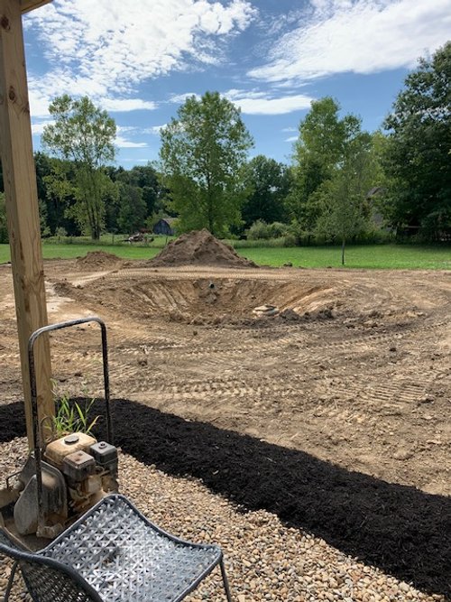 Backyard excavation site with a large hole and mounds of dirt, surrounded by trees and a grassy area, under a blue sky with clouds, and a metal structure in the foreground.