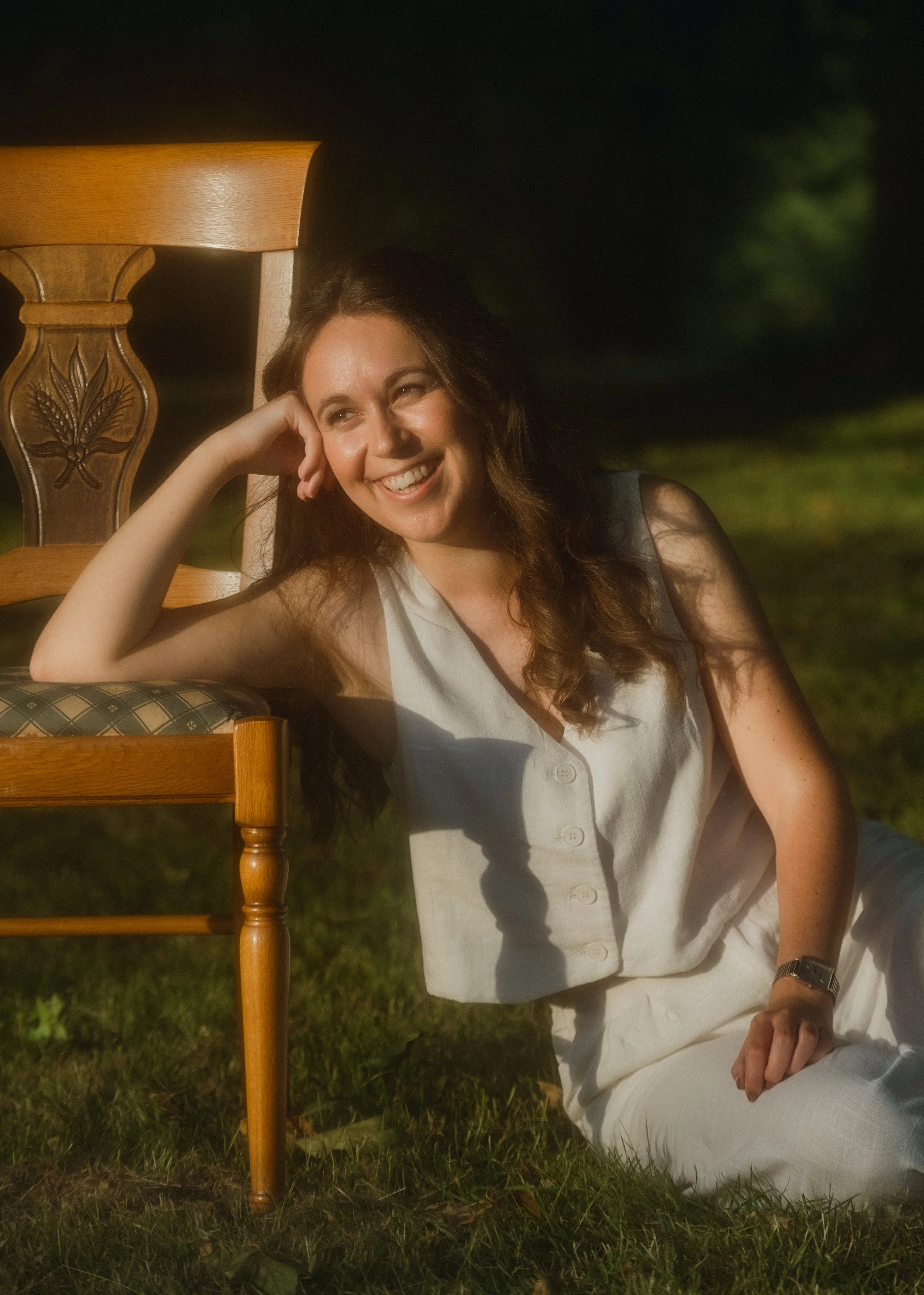 Une femme rieuse assise sur l'herbe à côté d'une chaise en bois en soirée.