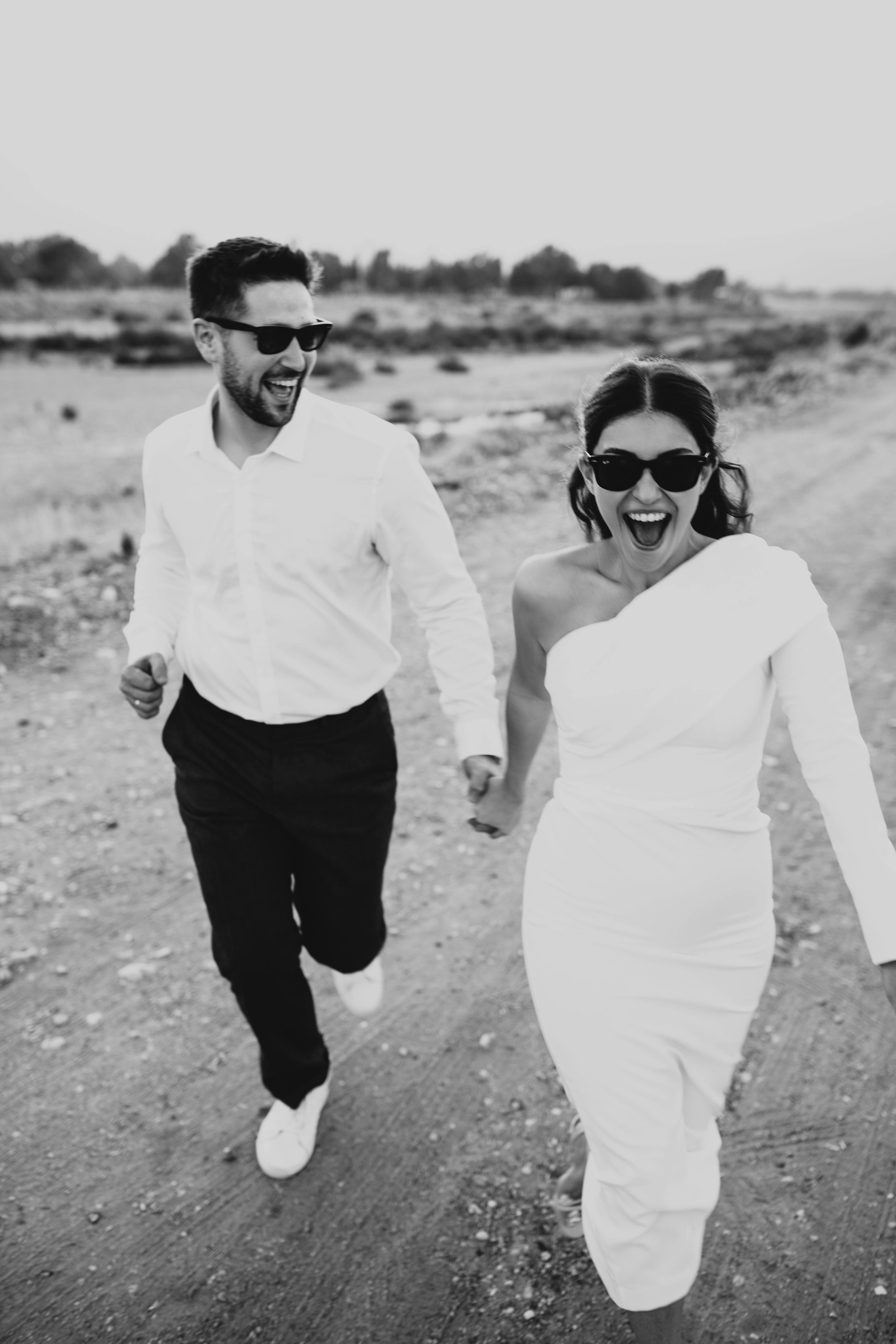 Un couple souriant et tenant la main en courant sur un chemin de terre, portant des lunettes de soleil et habillé élégamment, dans un paysage extérieur.