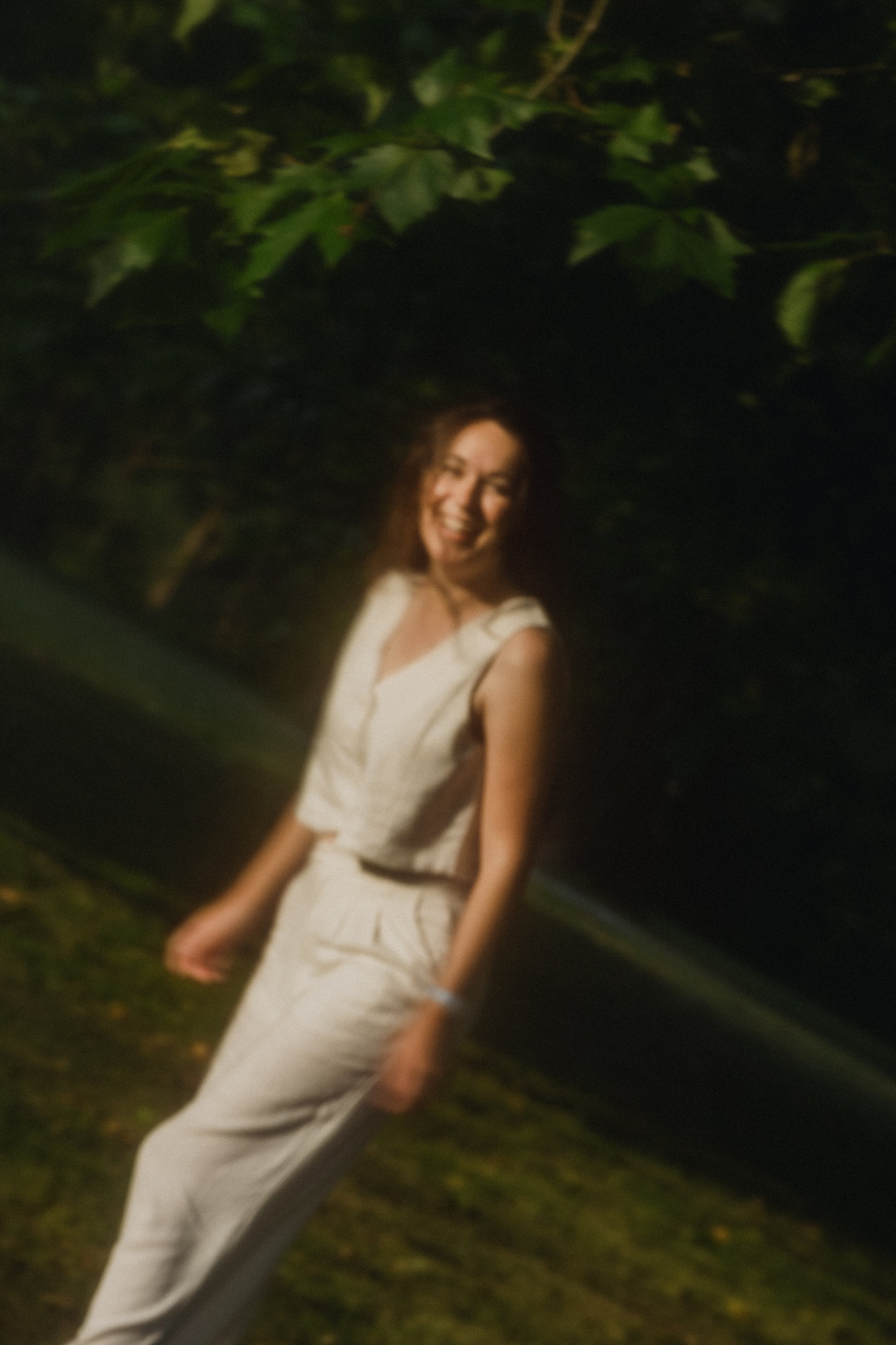 Une femme souriante portant une robe blanche dans un environnement extérieur avec des feuilles vertes.