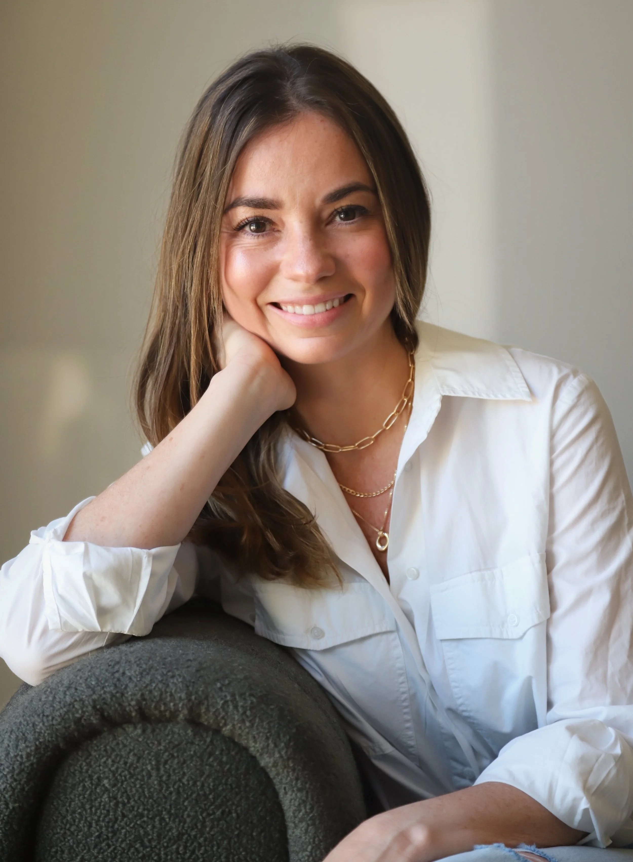 Retrato de una mujer joven con cabello castaño claro, sonriendo, usando camisa blanca y accesorios dorados, apoyada en un respaldo de sillón oscuro.