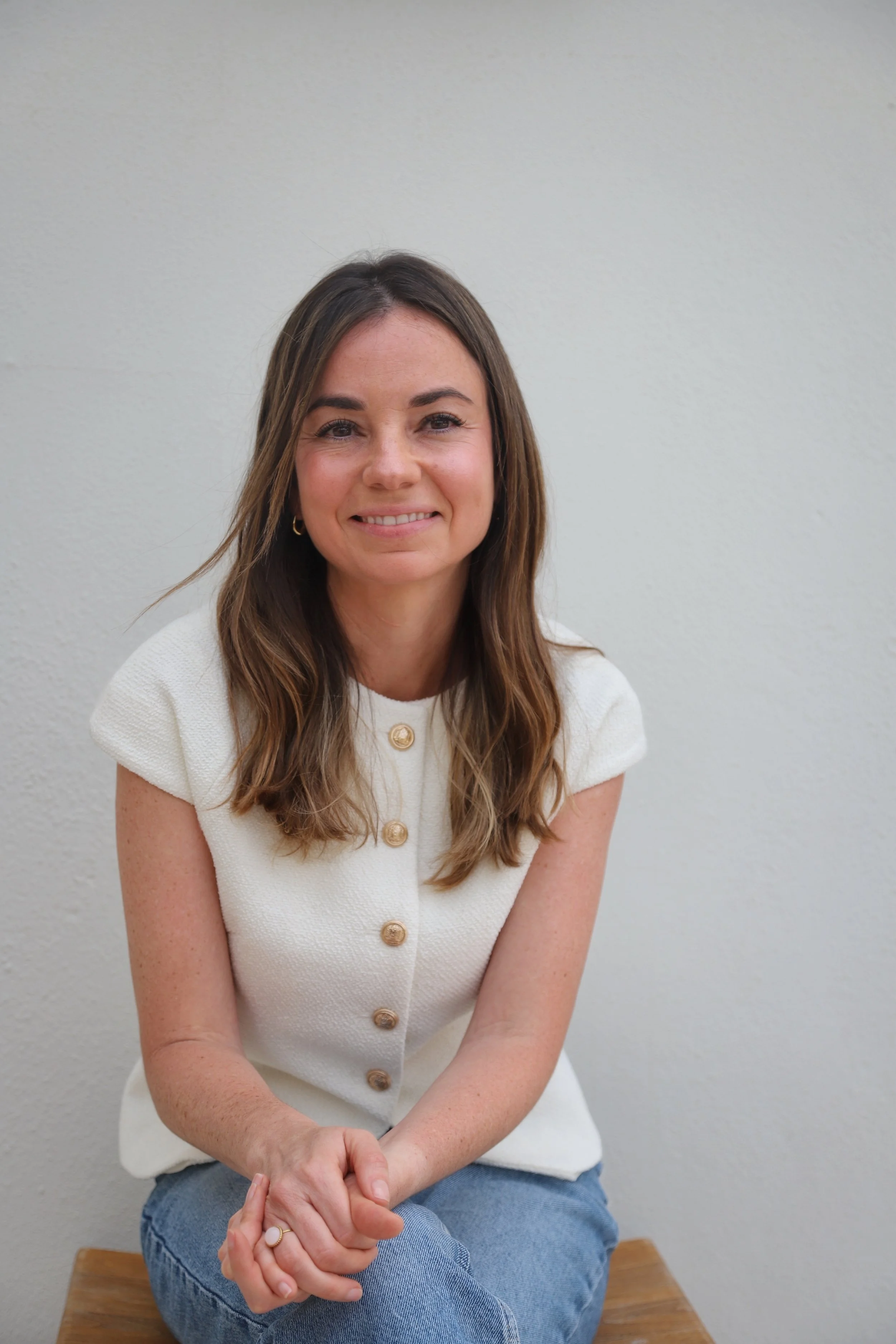 Mujer joven sonriente en silla de madera, con cabello castaño y vestido blanco con botones dorados, frente a pared blanca.