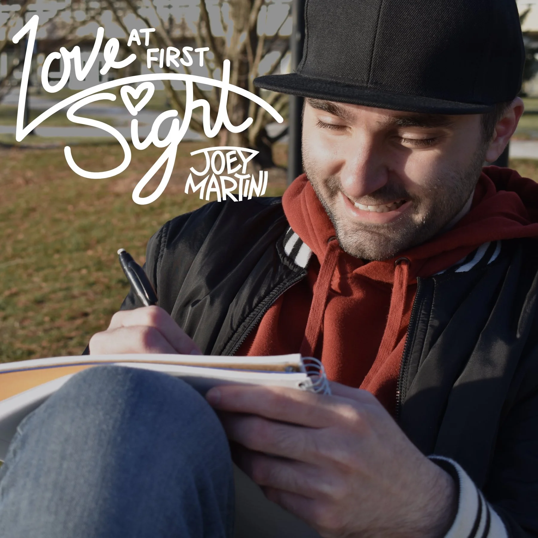 Love At First Sight single cover art featuring Joey smiling while writing in a notebook, with the song’s and Joey’s logo in the top left corner.