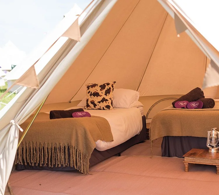 Deluxe bell tent interior with twin beds , soft bedding, lantern lighting, and wooden furniture creating a cosy boutique camping setup