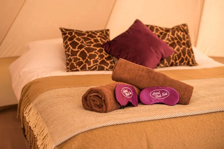 Luxury glamping tent bed with beige and white bedding, purple pillows, and soft rolled towels for a boutique camping stay.