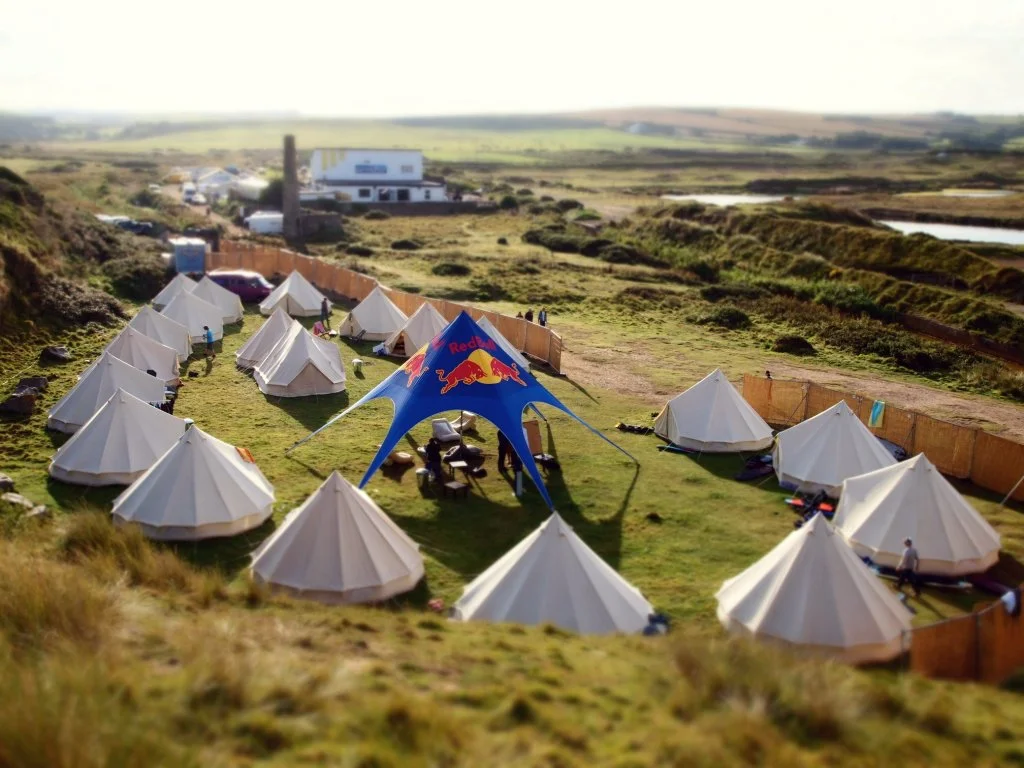 A campsite with white tents arranged in a semi-circle on a grassy field, featuring a large blue tent with a Red Bull logo in the center, with rolling hills and a body of water in the background.
