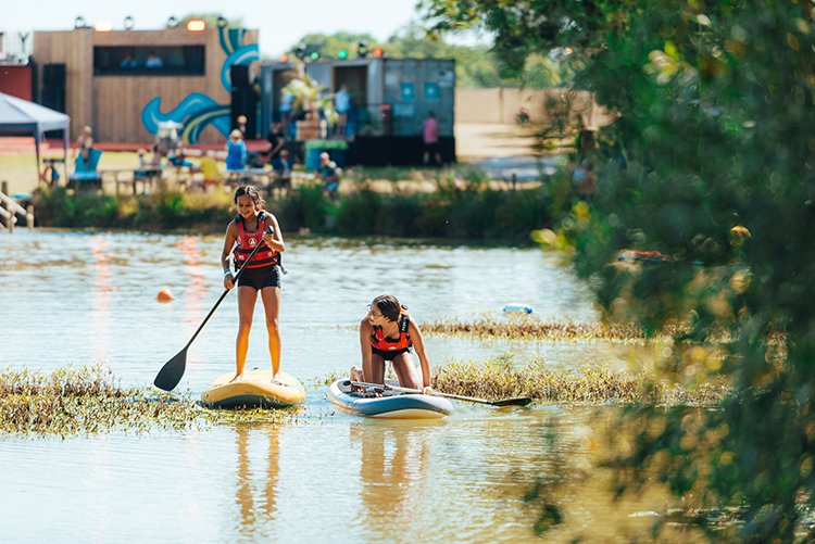 Two kids paddleboarding on a peaceful lake near a boutique glamping site surrounded by colourful festival scenery