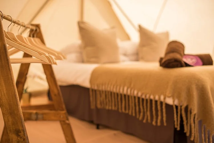 Cosy Super Bell tent interior with soft beige bedding, wooden clothes rail, and warm ambient lighting for a boutique camping stay